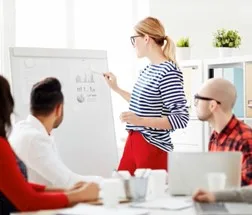 Drei Personen in einem hellen Büro, eine Frau an einem Whiteboard, zwei Männer am Tisch mit Laptops, die alle zusammenarbeiten. Dies zeigt IT Strategie-Beratung oder IT Strategy Consultant