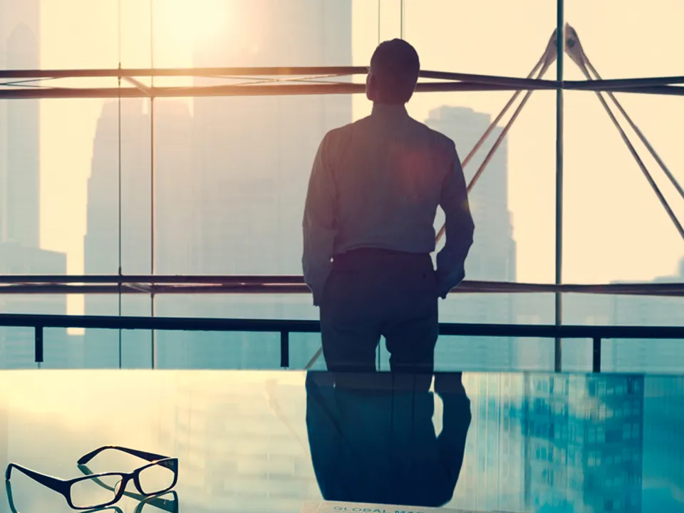 Person looking out of a window at a city skyline, a glass table with a pair of glasses in the foreground.